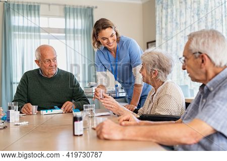 Nurse giving medicine to group of seniors at retirement community. Happy smiling nurse gives medicines to elderly patients at nursing home. Happy senior woman taking her dose of medicines.