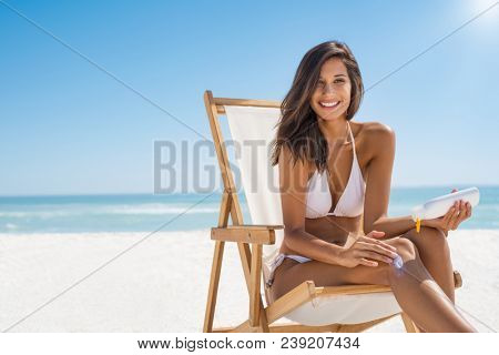 Young woman in bikini sitting on deckchair and applying sun lotion on leg. Portrait of smiling latin girl applying sun screen on body at beach with copy space. Beautiful woman enjoying sunbathing.