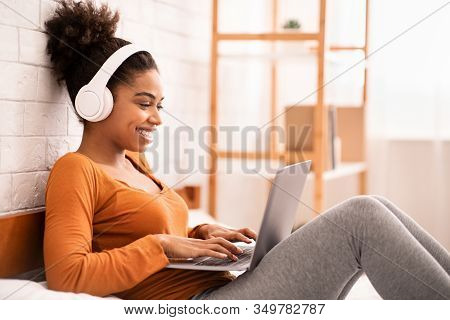 Smiling Afro Woman In Headphones Using Laptop Browsing Internet Relaxing Lying In Bed At Home. Weeke