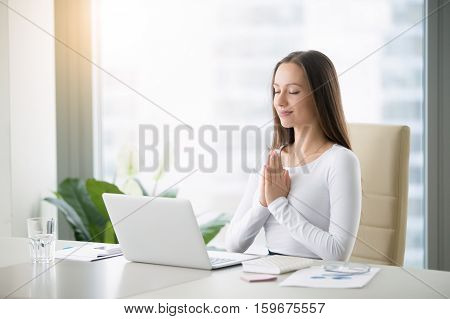 Young woman meditating sitting at the modern office desk in front of laptop, taking a pause, busy, stressful office, cure for work overload, one moment meditation, worshiping laptop