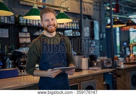 Successful small business owner using digital tablet and looking at camera. Happy smiling waiter ready to take order. Portrait of young entrepreneur of coffee shop standing at counter with copy space.