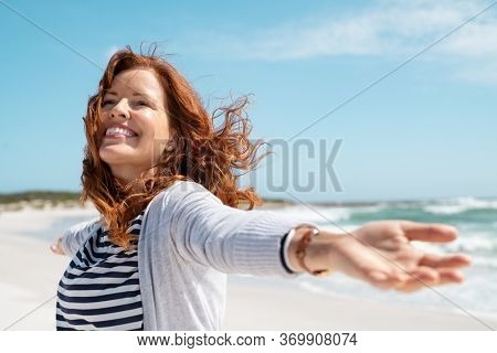 Happy mature woman with arms outstretched feeling the breeze at beach. Beautiful middle aged woman with arms up dancing on beach. Mid lady feeling good and enjoying freedom at sea, copy space.