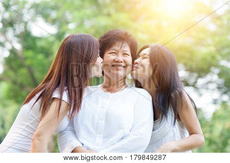 Portrait of Asian daughters kissing elderly mother, senior adult woman and grown child. Outdoors family at nature park with beautiful sun flare.