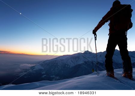 At dusk a brave backcountry skier reaching the summit of the mountain after a long day walking in the wilderness. Adventure and exploration concept.