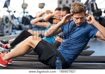 Muscular guy doing sit ups at gym with other people in background. Young athlete doing stomach workout in modern gym. Handsome fit man doing crunches at gym.