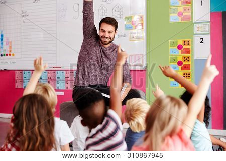 Elementary Pupils Raising Hands To Answer Question As Male Teacher Reads Story In Classroom