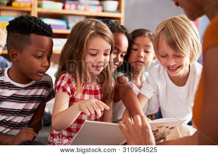 Group Of Elementary School Pupils Sitting On Floor Listening To Female Teacher Read Story