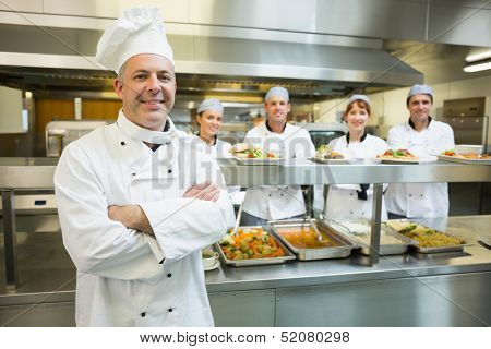 Proud mature head chef posing in a modern kitchen with his colleagues in the background