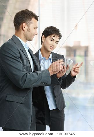 Due imprenditori in piedi nell'ufficio di lobby utilizzando smart mobile phone, sorridente.