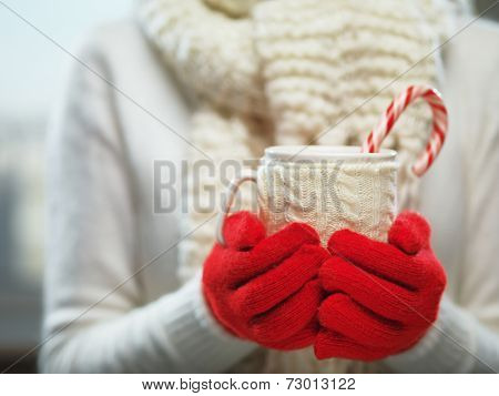 Woman holding winter cup close up on light background. Woman hands in woolen red gloves holding a cozy mug with hot cocoa, tea or coffee and a candy cane. Winter and Christmas time concept.