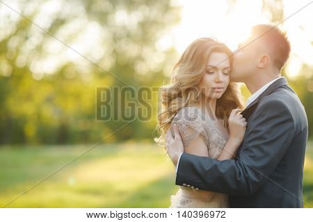 Young loving couple on the wedding day,the groom-a man with a short haircut,in a dark wedding suit and a beautiful bride-a woman with long curly hair in beige wedding dress standing arm in arm at sunset in summer green Park