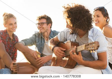 Happy friends having fun together while guy playing guitar. Group of young women and men enjoying their summer vacation. Cheerful young man playing guitar and his friends singing at sunset.