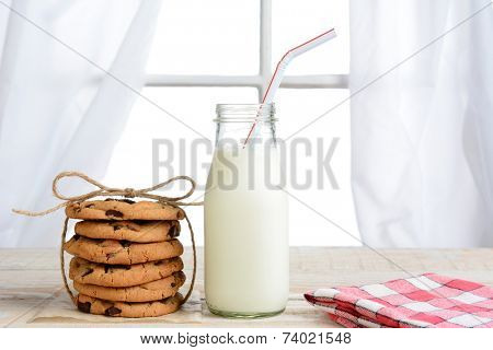 Horizontal shot of an after school snack of chocolate chip cookies and an old fashioned bottle of milk. The cookies are tied with twine and with a napkin on a rustic wood kitchen table.