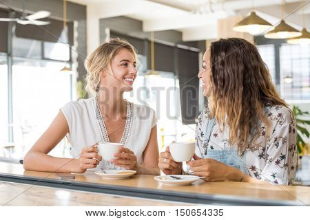 Two beautiful young women holding cup of coffee and talking to each other. Young blonde woman in conversation with her best friend while sipping a cup of capuccino. Friends meeting up for coffee.