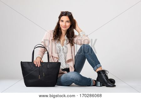 Cool young woman sitting down isolated on grey background. Young fashion woman looking at camera with trendy shopping bag. Stylish girl in casual clothes with accessory smiling.