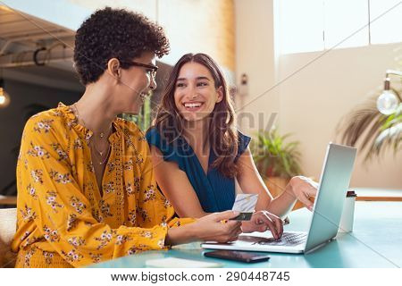 Happy woman making payment online using credit card. Cheerful friends discussing online shopping while purchasing products and paying bill amount using laptop and debit card. Two multiethnic girls.