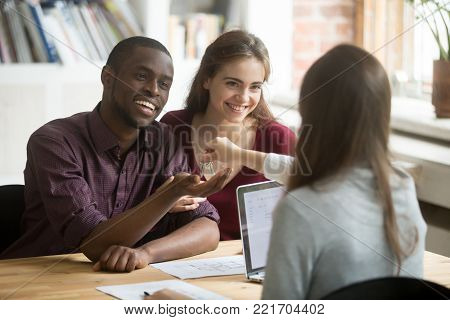 Happy multiracial couple getting keys to new home from realtor, excited young customers buying real estate house concept, diverse family buyers purchasing property, mortgage loan investment deal