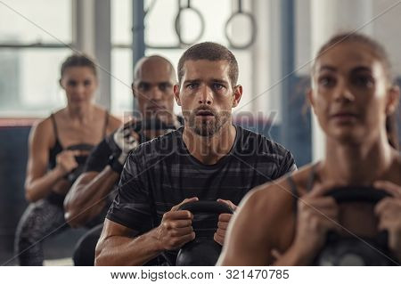 Portrait of fitness man lifting kettle bell with young women in fitness center. Active guy with group of people in gym doing weight lifting with kettlebell. Determined people in a row squatting.