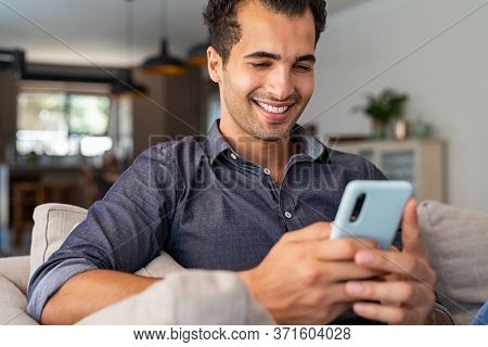 Cheerful businessman using smartphone while sitting on sofa at home. Handsome young indian man sitting on couch reading messages on mobile phone. Hispanic guy working from home with cellphone.