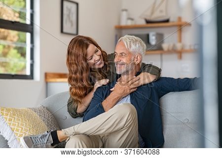 Smiling woman hugging her husband from behind in the living room. Loving mature couple sitting on couch and looking at each other. Mid adult beautiful woman embracing smiling senior at home.