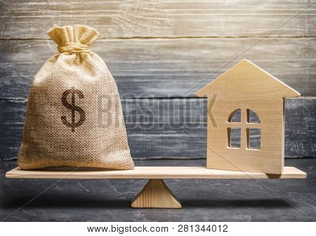 A Money Bag And A Wooden House On The Scales. The Concept Of Real Estate Purchase. Sale Of Property.