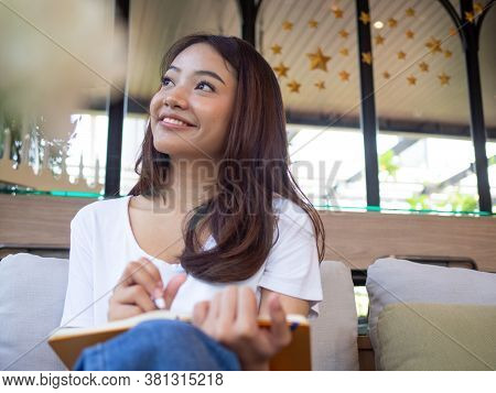 Young Asian Woman Or A Writer Sitting In A Journal Or Writing A Novel In A Coffee Shop Outside The O