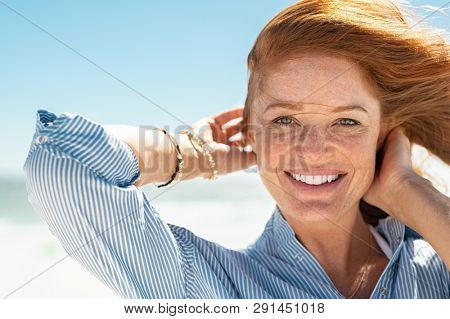 Portrait of beautiful mature woman with wind fluttering hair. Closeup face of healthy young woman with freckles looking at camera. Lady with red hair standing at seaside enjoying breeze at beach.