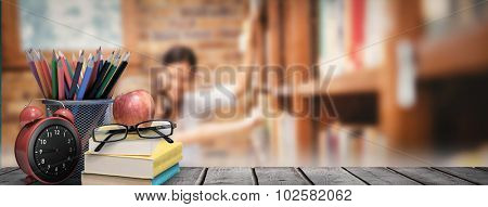 School supplies on desk against teacher and little girl selecting book in library