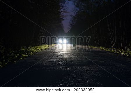 Male silhouette at the edge of a dark mountain road through the forest in the night. Scenic night landscape of dark blue sky. Man standing on the road against the light of car headlights. The car on the roadside.