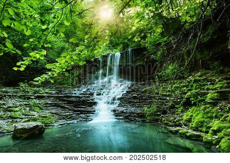 Beautiful mountain rainforest waterfall with fast flowing water and rocks, long exposure. Natural seasonal travel outdoor background with sun shihing