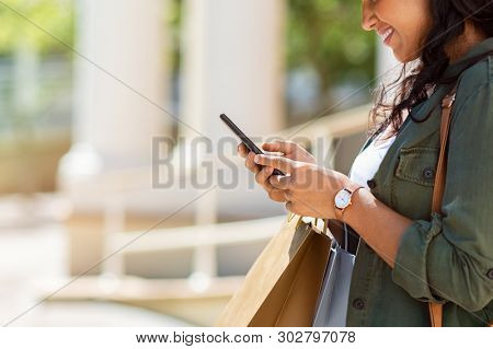 Closeup of woman hands holding shopping bags and using smartphone outdoor. Shopaholic girl checking online with smart phone shopping deals. Young stylish woman using mobile phone for ecommerce.