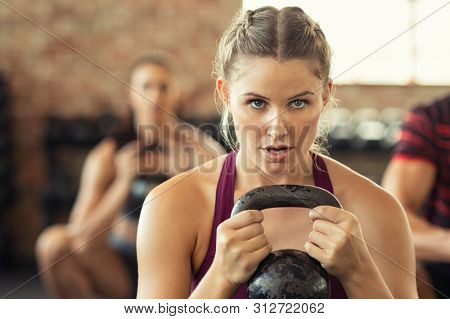 Portrait of concentrated young woman working out with kettlebell at gym. Closeup face of determined girl doing squat session and training biceps. Fitness class lifting heavy weights while squatting.