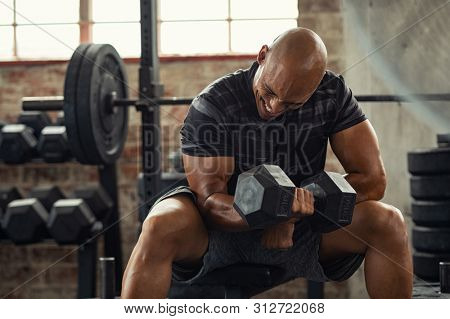 Muscular guy lifting dumbbell while sitting on bench at gym. Mature african athlete using dumbbell during workout. Strong man under physical exertion pumping up bicep muscle with heavy weight.