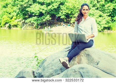 Young East Indian American Woman Wearing White Shirt, Black Pants, White Sneakers, Sitting On Rocks 