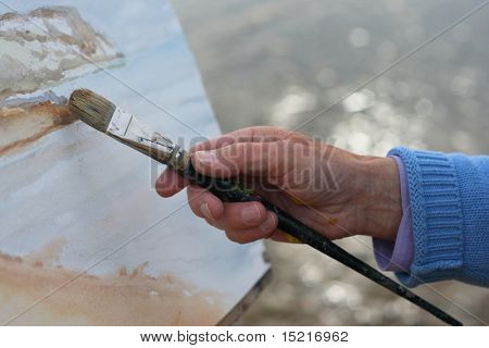 A close up of a mature artists hand painting a coastal landscape.