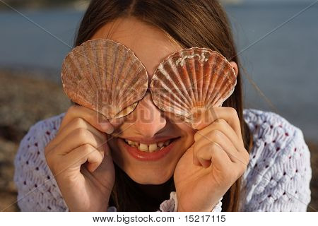 Young girl by the sea with seashells covering her eyes laughing.