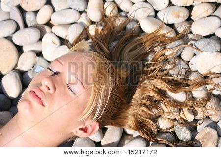 Young womans head at an angle with eyes shut lying on a pebble beach with wet hair spread.