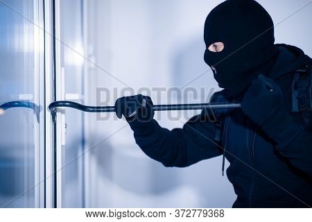 Intrusion Concept. Side View Portrait Of Disguised Prowler Breaking House Or Office Door With Crowba