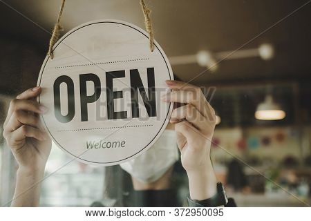 Open. Barista, Waitress Woman Wearing Protection Face Mask Turning Open Sign Board On Glass Door In 