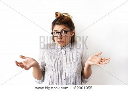 Indoor shot of emotional funny young woman in eyeglasses and formal shirt looking confused and puzzled her arms out shrugs shoulders. Emotions facial expressions feelings body language