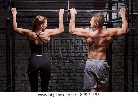 Rear view photo of muscular man and woman doing exercises on horizontal bar against brick wall at the cross fit gym. Sporty couple in the sportwear. Power and energy. Healthy lifestyle. Concept of the cross fit activity in the couple.