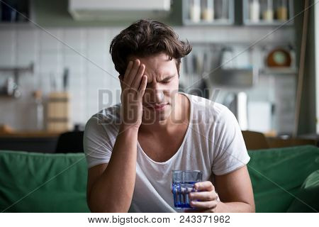 Young Man Suffering From Strong Headache Or Migraine Sitting With Glass Of Water In The Kitchen, Mil