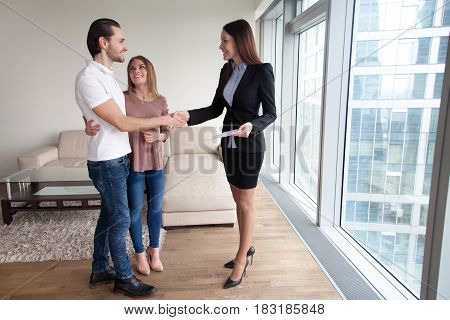 Happy beautiful couple and friendly realtor handshaking making real estate deal after signing sale purchase agreement or rental contract. Businesswoman and clients shaking hands, full length portrait