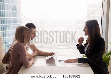 Side view of realtor and young couple sitting at office desk discussing property for sale. Potential buyers holding house plan, considering investment in home or taking loan to purchase real estate