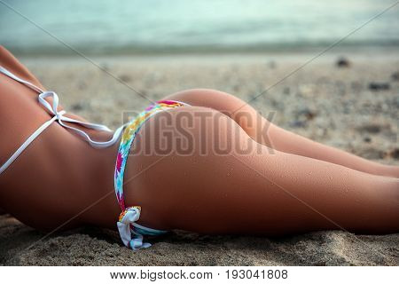 Sporty young beautiful woman in bikini sunbathing with a sandy sexy butt on the sea shore. Outdoor lifestyle picture on a hot sunny summer day. Sexy buttocks