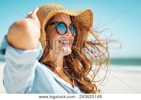 Side view of beautiful mature woman wearing sunglasses at beach. Young smiling woman on vacation looking away while enjoying sea breeze wearing straw hat. Closeup portrait of attractive girl relaxing.