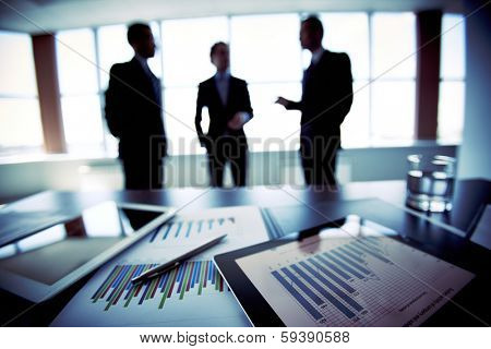 Close-shot of a tablet computer displaying financial data, three businessmen standing in the background