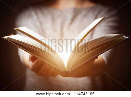 Light coming from book in woman's hands in gesture of giving, offering. Concept of wisdom, religion, reading, imagination.