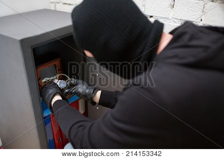 theft, burglary and people concept - thief in mask stealing valuables from safe at crime scene