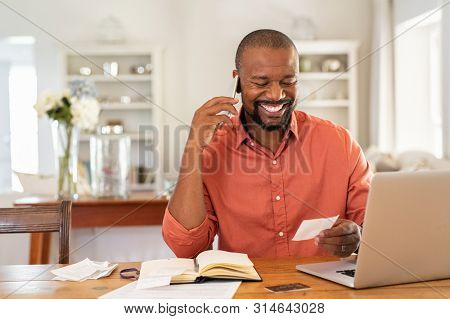 Smiling african man using laptop at home while checking home finance. Happy mature man looking at invoice while talking on phone with bank. Man checking receipt and bill while talking at phone.